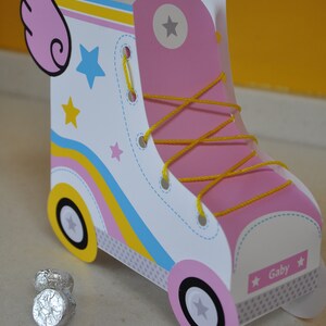 Peut inclure: Une bo&icirc;te de faveur de f&ecirc;te en forme de patin &agrave; roulettes rose et blanche avec des lacets jaunes et une bande bleue et jaune. La bo&icirc;te a une aile rose sur le c&ocirc;t&eacute; et dit "Gaby" sur le devant.