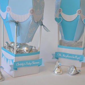 May include: Two blue and white hot air balloon favor boxes with white and blue striped bases. The boxes are filled with silver foil-wrapped chocolates. One box has a banner that reads "Oh, the places he'll go!" and the other box has a banner that reads "Christy's Baby Shower".