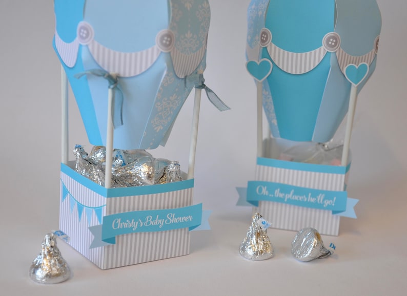 May include: Two blue and white hot air balloon favor boxes with white and blue striped bases. The boxes are filled with silver foil-wrapped chocolates. One box has a banner that reads "Oh, the places he'll go!" and the other box has a banner that reads "Christy's Baby Shower".