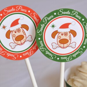 May include: Two round, festive cupcake toppers with a dog wearing a Santa hat and holding a bone. The toppers are red and green with the text "Santa Paws" repeated around the edge.