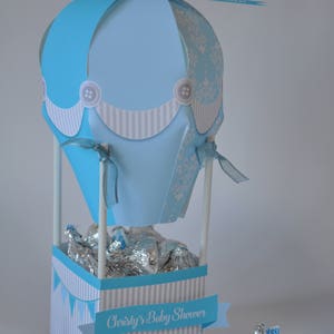 May include: A blue and white hot air balloon shaped party favor box with a banner that reads "Christy's Baby Shower". The box is filled with silver foil wrapped chocolates.