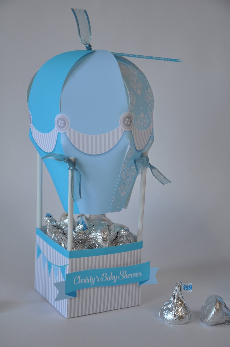 May include: A blue and white hot air balloon shaped party favor box with a banner that reads "Christy's Baby Shower". The box is filled with silver foil wrapped chocolates.