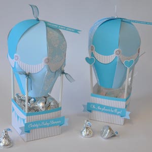 May include: Two blue and white hot air balloon centerpieces with silver foil-wrapped chocolate kisses inside. The balloons are decorated with white buttons and a white and blue striped border. The centerpieces are labeled "Welcome Baby Boy" and "Oh, the places he'll go!"
