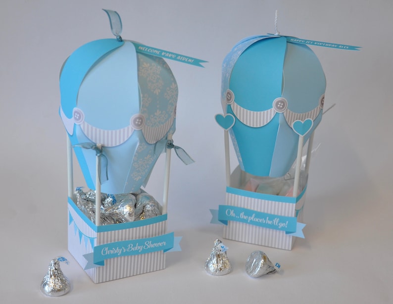 May include: Two blue and white hot air balloon centerpieces with silver foil-wrapped chocolate kisses inside. The balloons are decorated with white buttons and a white and blue striped border. The centerpieces are labeled "Welcome Baby Boy" and "Oh, the places he'll go!"