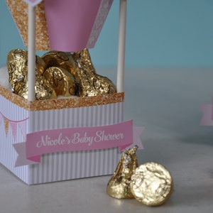 May include: Pink and gold hot air balloon favour boxes filled with chocolate kisses. The boxes have a white and grey striped pattern and a pink banner that says "Nicole's Baby Shower".