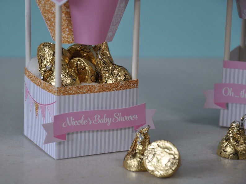 May include: Pink and gold hot air balloon favour boxes filled with chocolate kisses. The boxes have a white and grey striped pattern and a pink banner that says "Nicole's Baby Shower".