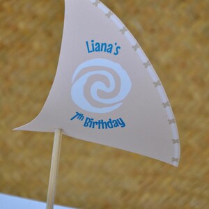 May include: A beige paper sail with a blue and white swirl design and the text "Liana's 7th Birthday". The sail is attached to a wooden stick and is part of a party decoration.
