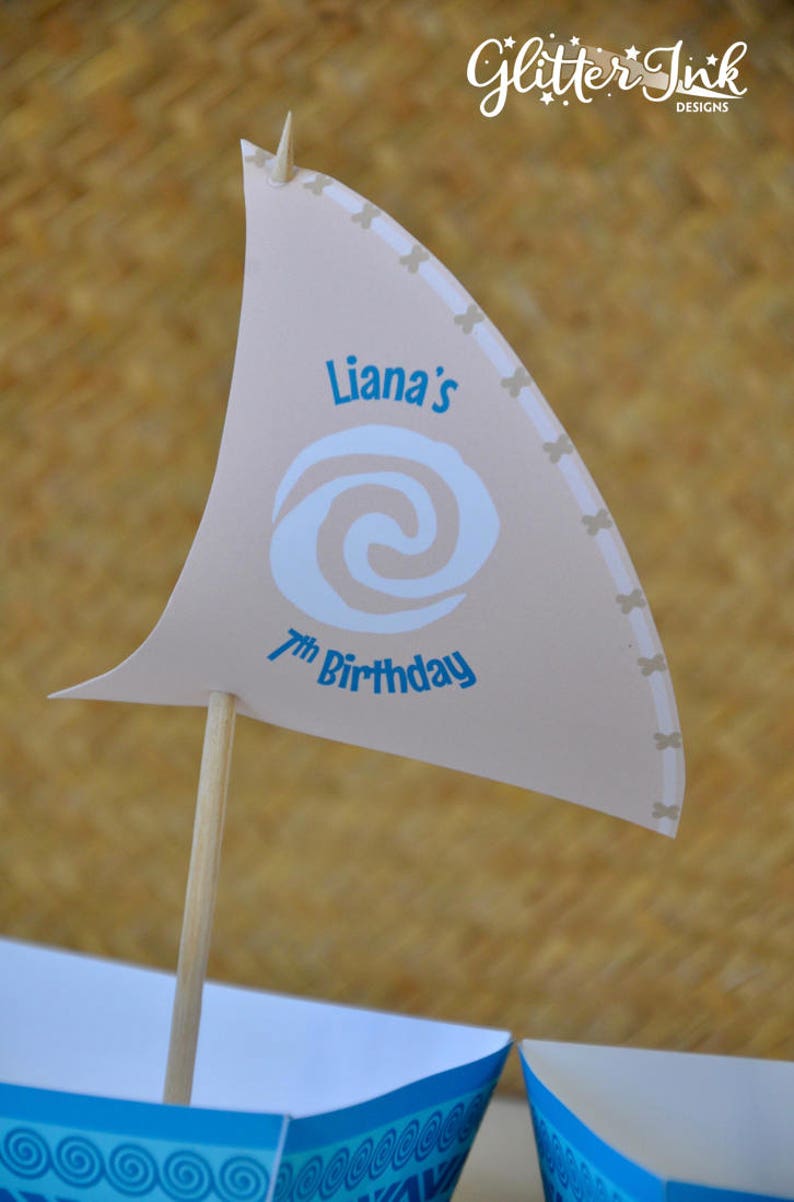 May include: A beige paper sail with a blue and white swirl design and the text "Liana's 7th Birthday". The sail is attached to a wooden stick and is part of a party decoration.