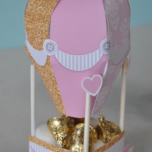 May include: A pink and gold hot air balloon party favour box with a white and grey striped banner and a heart-shaped cutout. The box is filled with chocolate kisses and has a pink banner that says "Nicole's Baby Shower".
