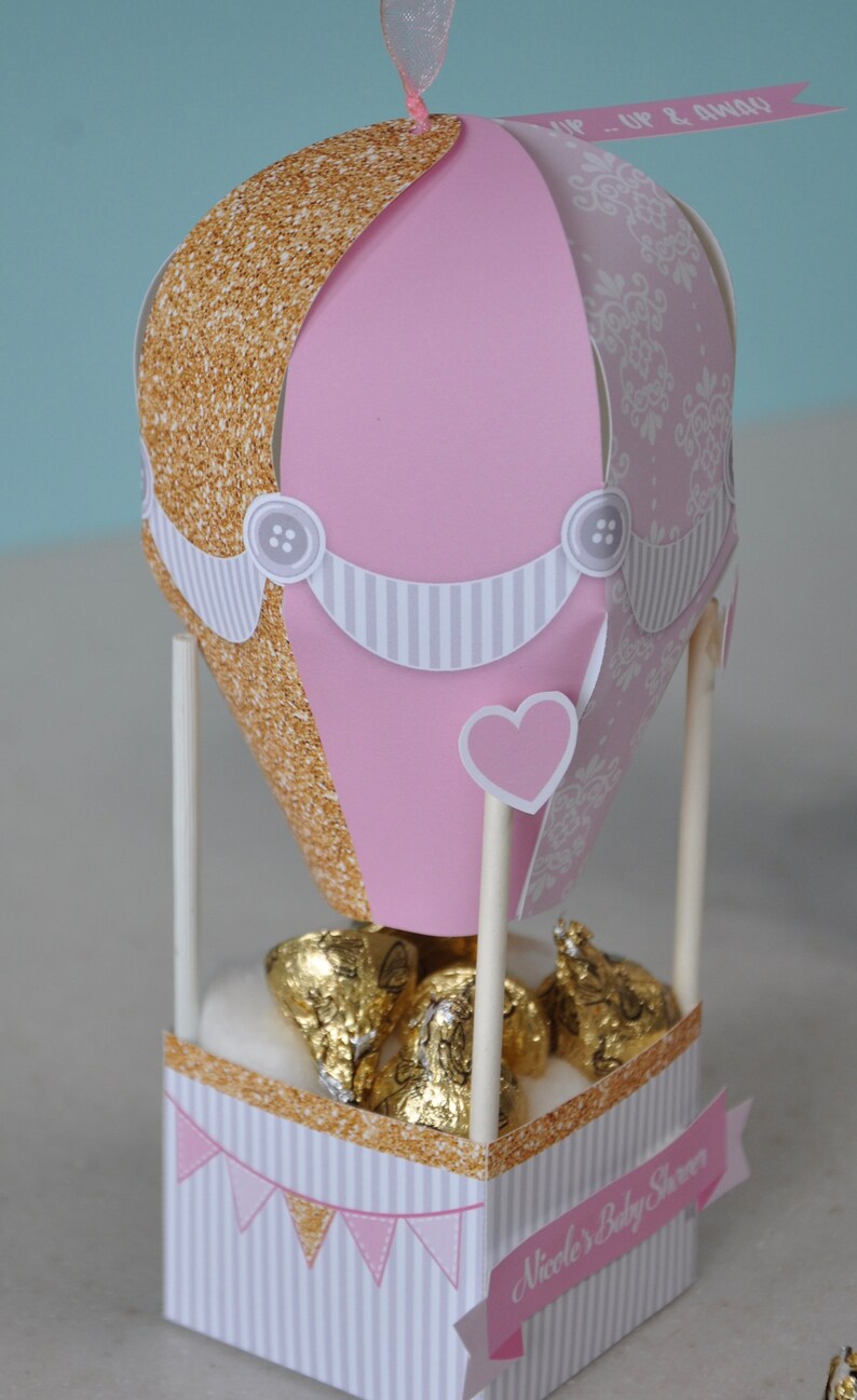 May include: A pink and gold hot air balloon party favour box with a white and grey striped banner and a heart-shaped cutout. The box is filled with chocolate kisses and has a pink banner that says "Nicole's Baby Shower".