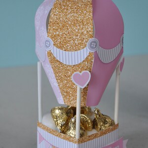 May include: Pink and gold hot air balloon shaped favour box with white stripes and a heart design. The box is filled with chocolate kisses and has a banner that says "Nicole's Baby Shower".