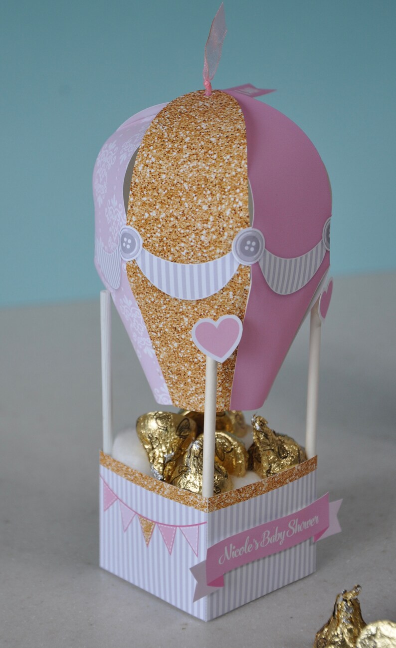May include: Pink and gold hot air balloon shaped favour box with white stripes and a heart design. The box is filled with chocolate kisses and has a banner that says "Nicole's Baby Shower".