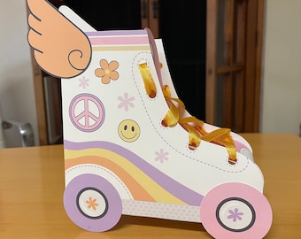 Può includere: Una scatola di favore per feste in cartone a forma di pattino a rotelle, di colore bianco e viola, con un nastro giallo, un segno di pace, un fiore e decorazioni a forma di smiley. La scatola è a forma di pattino a rotelle con ruote.