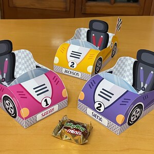 以下が含まれることがあります： チェック柄の旗が付いた、ピンク、イエロー、パープルの3つの段ボール製レーシングカーのギフトボックス。各ボックスには番号、名前、3本の黒いストライプがあります。ボックスは、チョコレートバーのある木製の表面の上に置かれています。