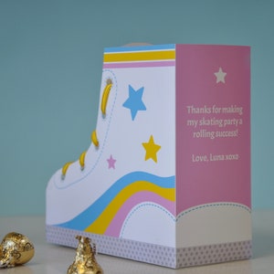 Peut inclure: Une bo&icirc;te &agrave; drag&eacute;es en carton blanc en forme de patin &agrave; roulettes avec des rayures jaunes, bleues et roses. La bo&icirc;te contient un message de remerciement qui dit "Merci d'avoir fait de ma f&ecirc;te de patinage un succ&egrave;s retentissant ! Avec amour, Luna xoxo".