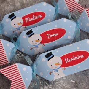 Puede incluir: Tres crackers navideños azules y rojos con ilustraciones de muñecos de nieve y los nombres "Madison", "Drem" y "Verónica" impresos en ellos.