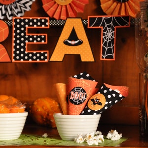 May include: A Halloween party treat table with orange and black decorations, including a banner that reads "Treat", and a pumpkin with a friendly face. There are also small cones filled with popcorn and candy.