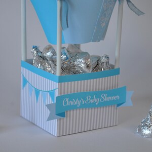 May include: A blue and white striped box with a blue banner that says "Christy's Baby Shower". The box is filled with silver foil-wrapped chocolate kisses.