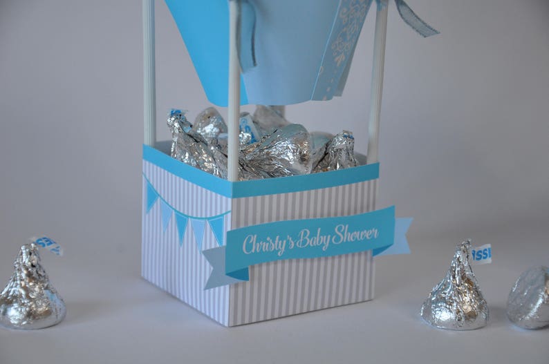 May include: A blue and white striped box with a blue banner that says "Christy's Baby Shower". The box is filled with silver foil-wrapped chocolate kisses.