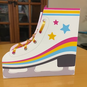 May include: A white cardboard ice skate party favor box with a yellow ribbon, pink, yellow, and blue stripes, and gold and blue stars.