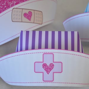 Op de afbeelding: Drie witte papieren verpleegstersmutsen met roze rand. De mutsen hebben verschillende ontwerpen: een roze pleister met een hart, paarse strepen en een roze kruis met een hart.