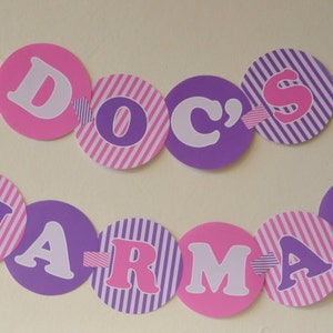 May include: A pink and purple paper banner with the words "Doc's Pharmacy" in white letters. The banner is strung together with white string and has small pink hearts and bandages attached to it.