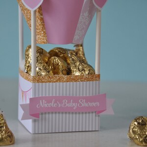 May include: Pink and white hot air balloon shaped favour boxes filled with gold foil wrapped chocolate kisses. The boxes have a gold glitter trim and a pink banner that reads "Nicole's Baby Shower".