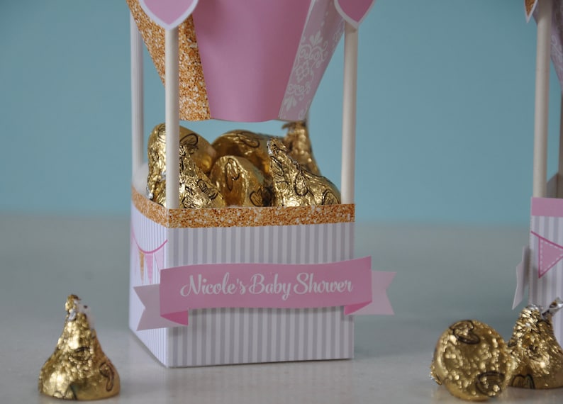 May include: Pink and white hot air balloon shaped favour boxes filled with gold foil wrapped chocolate kisses. The boxes have a gold glitter trim and a pink banner that reads "Nicole's Baby Shower".