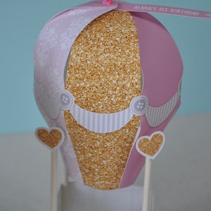 May include: A pink and gold hot air balloon centrepiece with a white and grey striped base. The balloon has a gold glitter pattern and a white lace design. The balloon has a banner that says "Alana's 1st Birthday".