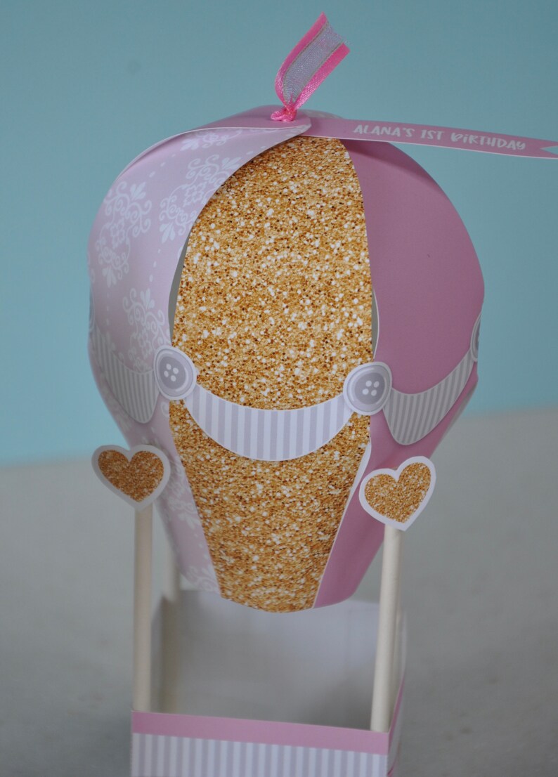 May include: A pink and gold hot air balloon centrepiece with a white and grey striped base. The balloon has a gold glitter pattern and a white lace design. The balloon has a banner that says "Alana's 1st Birthday".