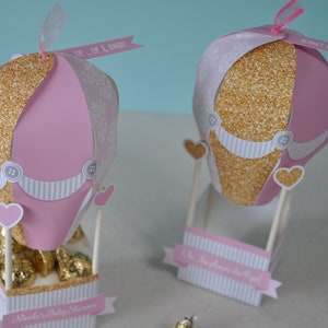 May include: Two pink and gold hot air balloon party favours with white and grey striped details. The balloons are decorated with glitter, hearts, and buttons. One balloon has a tag that says "Up, Up & Away" and the other says "Alana's 1st Birthday".