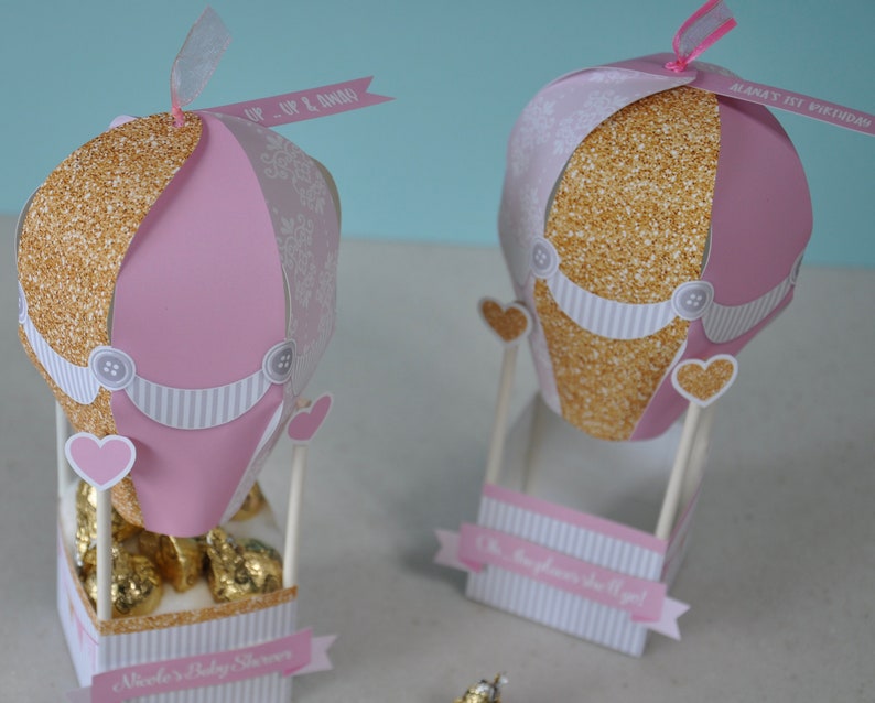 May include: Two pink and gold hot air balloon party favours with white and grey striped details. The balloons are decorated with glitter, hearts, and buttons. One balloon has a tag that says "Up, Up & Away" and the other says "Alana's 1st Birthday".