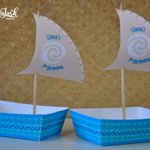 May include: Two blue and white paper party favor boxes with a sail design. The sails are beige and have a blue swirl design with the text "Liana's 7th Birthday".