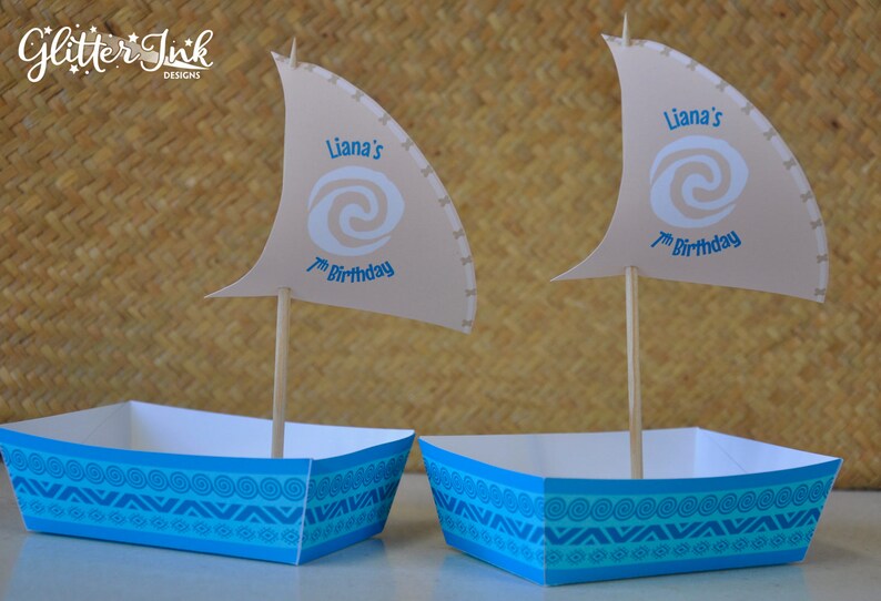 May include: Two blue and white paper party favor boxes with a sail design. The sails are beige and have a blue swirl design with the text "Liana's 7th Birthday".
