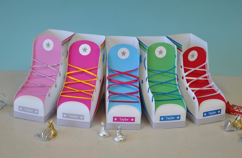Peut inclure: Cinq bo&icirc;tes &agrave; chaussures en papier color&eacute;es avec des lacets blancs et une &eacute;toile sur le c&ocirc;t&eacute;. Chaque bo&icirc;te porte le nom "Taylor" imprim&eacute; sur le fond. Les bo&icirc;tes sont pos&eacute;es sur une surface gris clair.