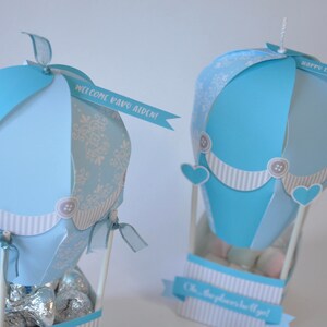 May include: Two blue and white hot air balloon party favors with ribbons and personalized messages. The balloon on the left says "Welcome Baby Aiden!" and the balloon on the right says "Happy 1st Birthday Alex!"