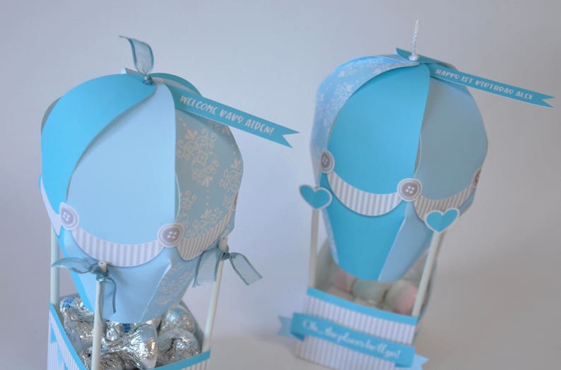 May include: Two blue and white hot air balloon party favors with ribbons and personalized messages. The balloon on the left says "Welcome Baby Aiden!" and the balloon on the right says "Happy 1st Birthday Alex!"