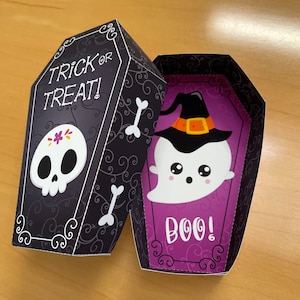 Könnte beinhalten: Eine schwarze und violette Pappbox in Sargform mit einem Totenkopf- und Knochenkreuz-Design und dem Text "Trick or Treat!" auf der Außenseite. Die Innenseite der Box hat einen violetten Hintergrund mit einem weißen Geist, der einen schwarzen Hexenhut trägt, und dem Text "Boo!"