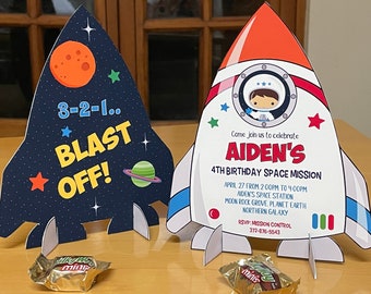 May include: Two rocket-shaped birthday invitations. One is navy blue with a planet and the words "3-2-1... BLAST OFF!". The other is white with an astronaut and the text "AIDEN'S 4TH BIRTHDAY SPACE MISSION".