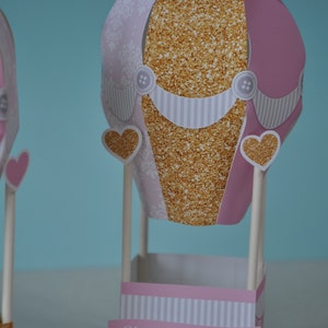 May include: A pink and gold hot air balloon party favour box with a white striped base and the text "Oh... the places she'll go!" printed on a banner.