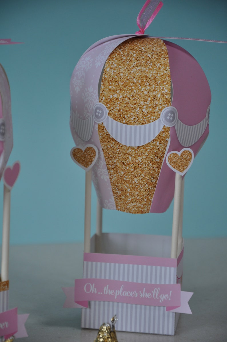 May include: A pink and gold hot air balloon party favour box with a white striped base and the text "Oh... the places she'll go!" printed on a banner.