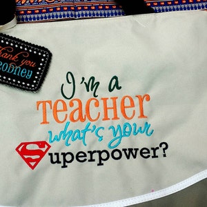 May include: A white canvas tote bag with a gray bottom and a white trim. The bag has an embroidered design that says "I'm a teacher what's your superpower?" with a red Superman logo. A black tag with white stitching says "Thank you Ms Rodney".