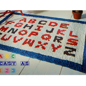 May include: A white rug with a blue border and red and black letters spelling out the alphabet. The rug is on a wooden floor with a red chair and a potted plant in the background. The text "ABC EASY AS 123" is printed on the floor below the rug.