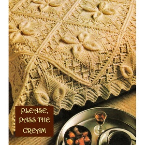 May include: A close-up of a crocheted blanket with a delicate pattern of flowers and bows. The blanket is a light cream color and is draped over a brown surface. The text "Please, pass the cream" is printed on a brown rectangle in the lower left corner of the image.