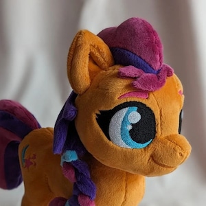 Könnte beinhalten: Ein Plüschpferd in Orange-, Lila- und Rosatönen. Das Pony hat eine geflochtene Mähne, einen Hut und große blaue Augen. Die Hufe sind weiß und rosa. Das Spielzeug steht auf einer hellen Oberfläche.