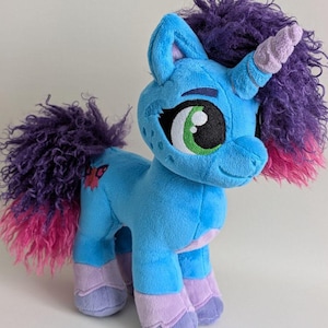 Könnte beinhalten: Ein Plüsch-Einhorn in Blau-, Lila- und Rosatönen. Das Einhorn hat einen blauen Körper, eine lila Mähne und einen Schweif mit rosa Spitzen und lavendelfarbene Hufe. Es hat ein grün-schwarzes Auge und einen kleinen Schmetterling an der Flanke.