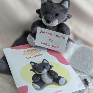 May include: A grey wolf plush toy holding a sign that says "Wanna Learn to Make Me?"  The plush toy is sitting on a white surface next to a book titled "Lobito the little wolf Sewpoke" by Zoe White.