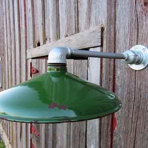 May include: A green metal barn light fixture with a round shade mounted on a weathered wooden barn wall. The light fixture is made of industrial pipe and has a silver finish.