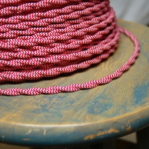 May include: A close-up of a spool of red and white braided electrical cord. The cord is wound tightly around the spool and has a zig-zag pattern.