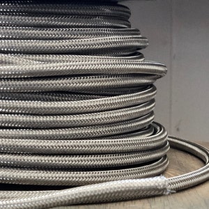 May include: A spool of silver braided wire, wound tightly around a black plastic core. The wire is flexible and has a smooth, braided texture.